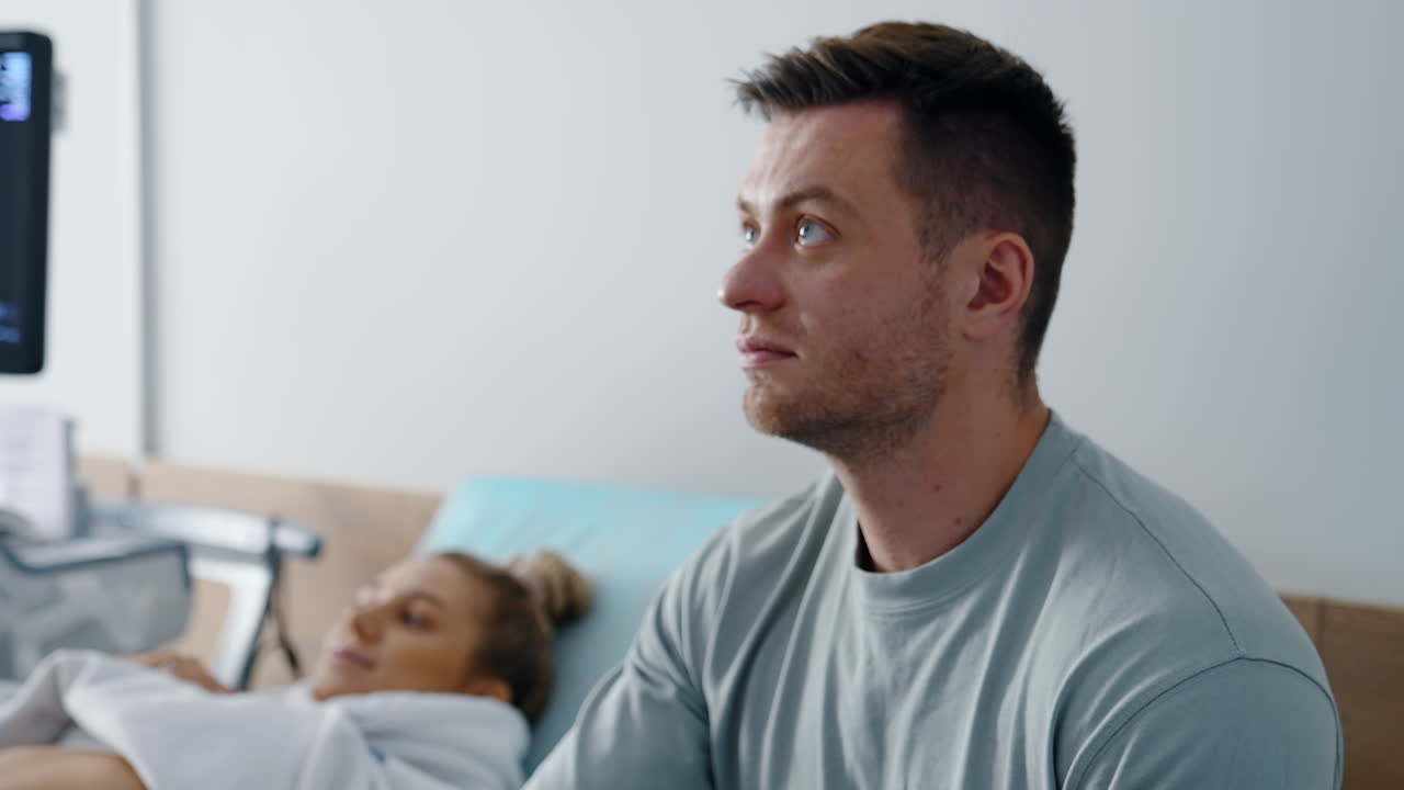 Focused bearded man looks up at the screen showing his future baby. Blonde woman lies on the couch at backdrop in blur.