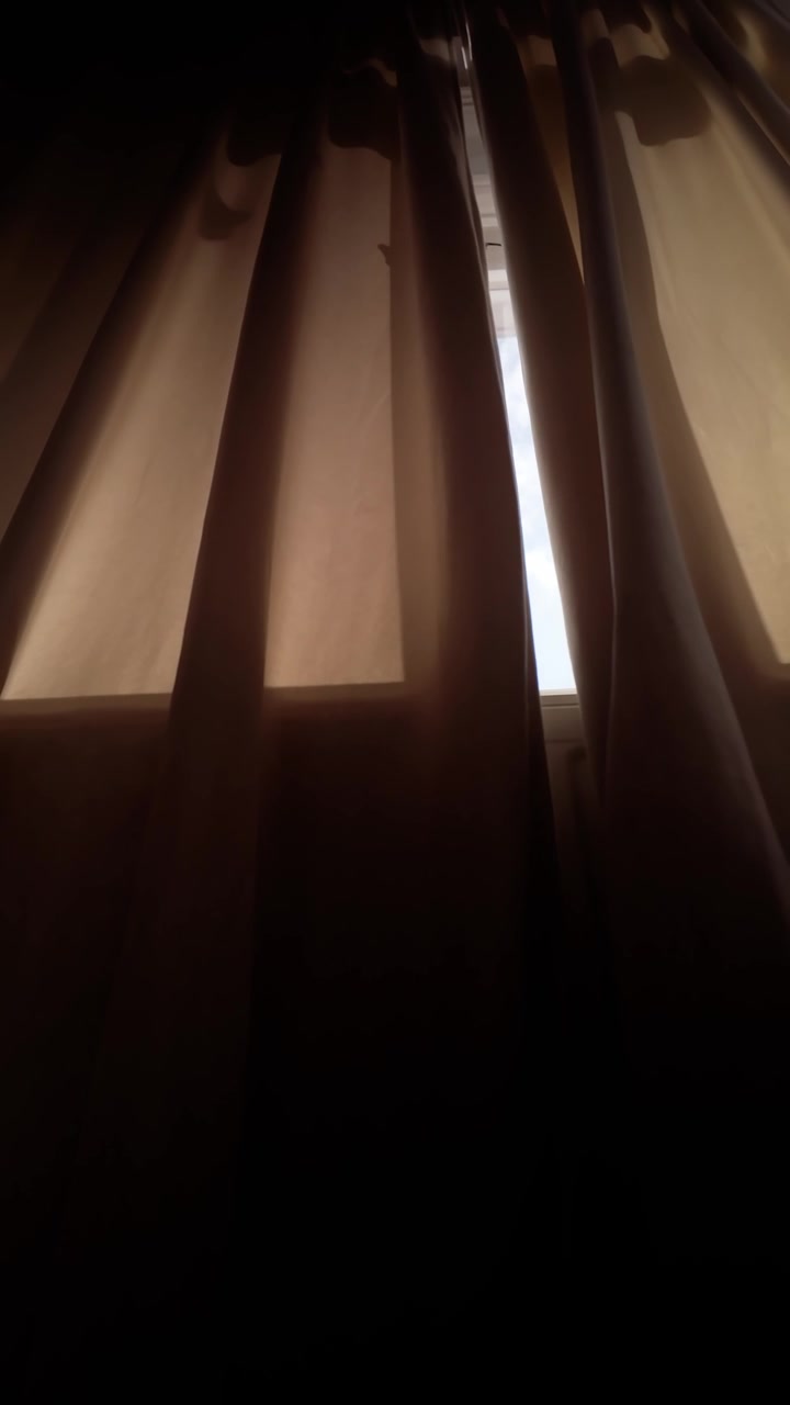 Vertical low angle view of morning light appearing through a narrow gap between curtains in a darkened bedroom, evoking a new day and daily ritual.