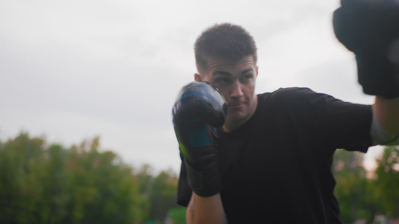 Boxer maschio bianco in un parco che alza i guantoni in posizione di guardia, postura concentrata, guantoni neri, guanti da allenatore nelle vicinanze, cielo nuvoloso, esercizi di difesa ripetitivi, gioco di gambe controllato e respirazione costante