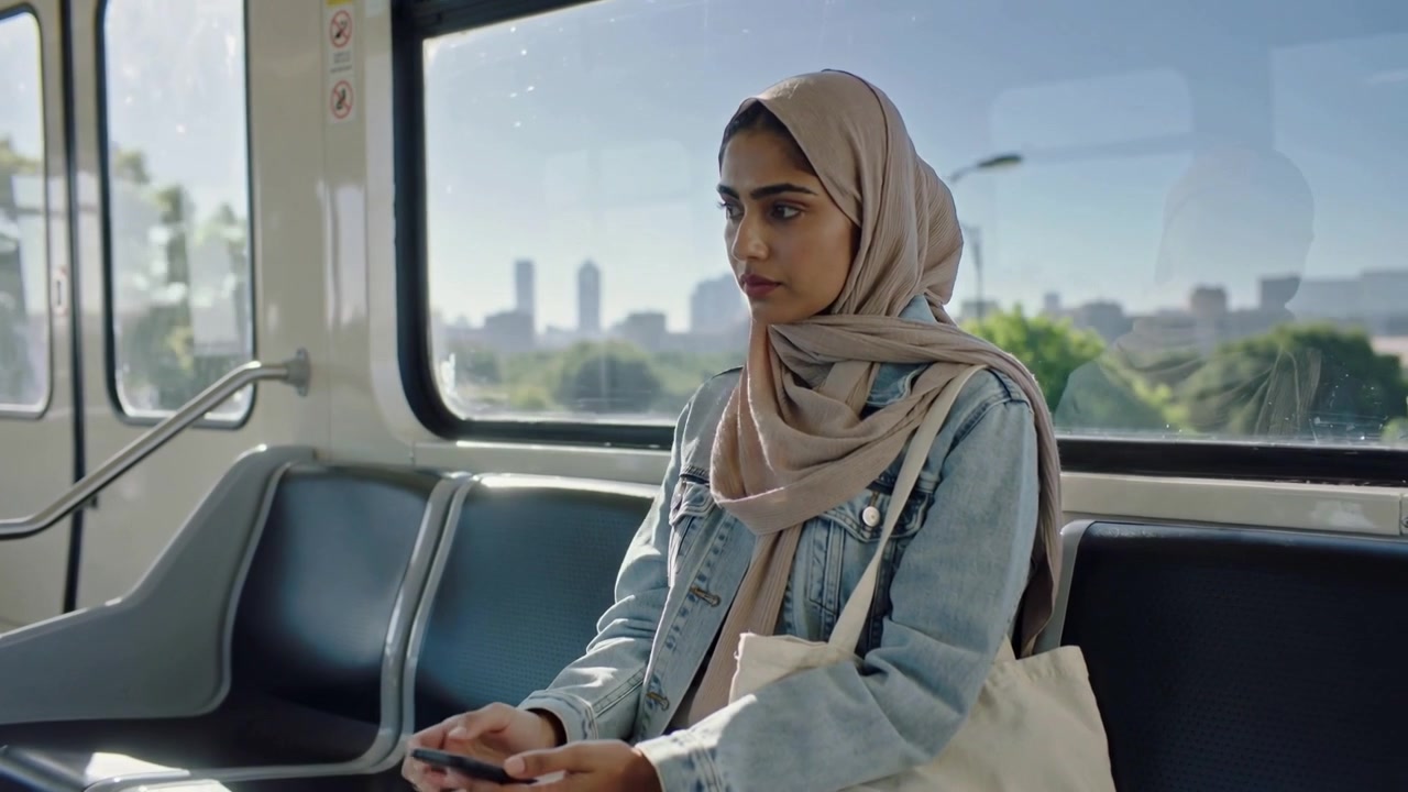 Una donna con hijab beige, vedendo lo smartphone, sistema l'auricolare sul treno leggero per ascoltare; una borsa tote è visibile.