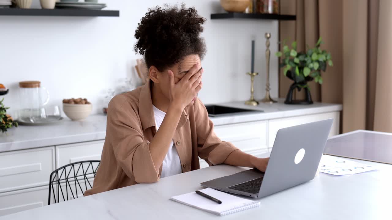 frustrada molesta afroamericana chica morena, independiente, gerente, se sienta en la mesa en la cocina, utiliza la computadora portátil mientras trabaja a distancia, conmocionada por malas noticias o mensajes, sufrió pérdidas financieras, desesperación