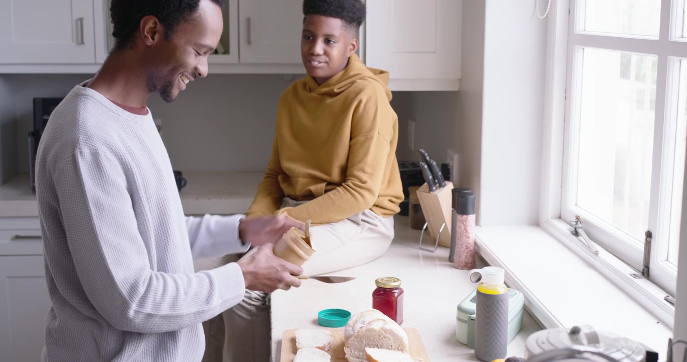 African American dad at counter holding peanut butter jar to teen, spreading on bread making snack