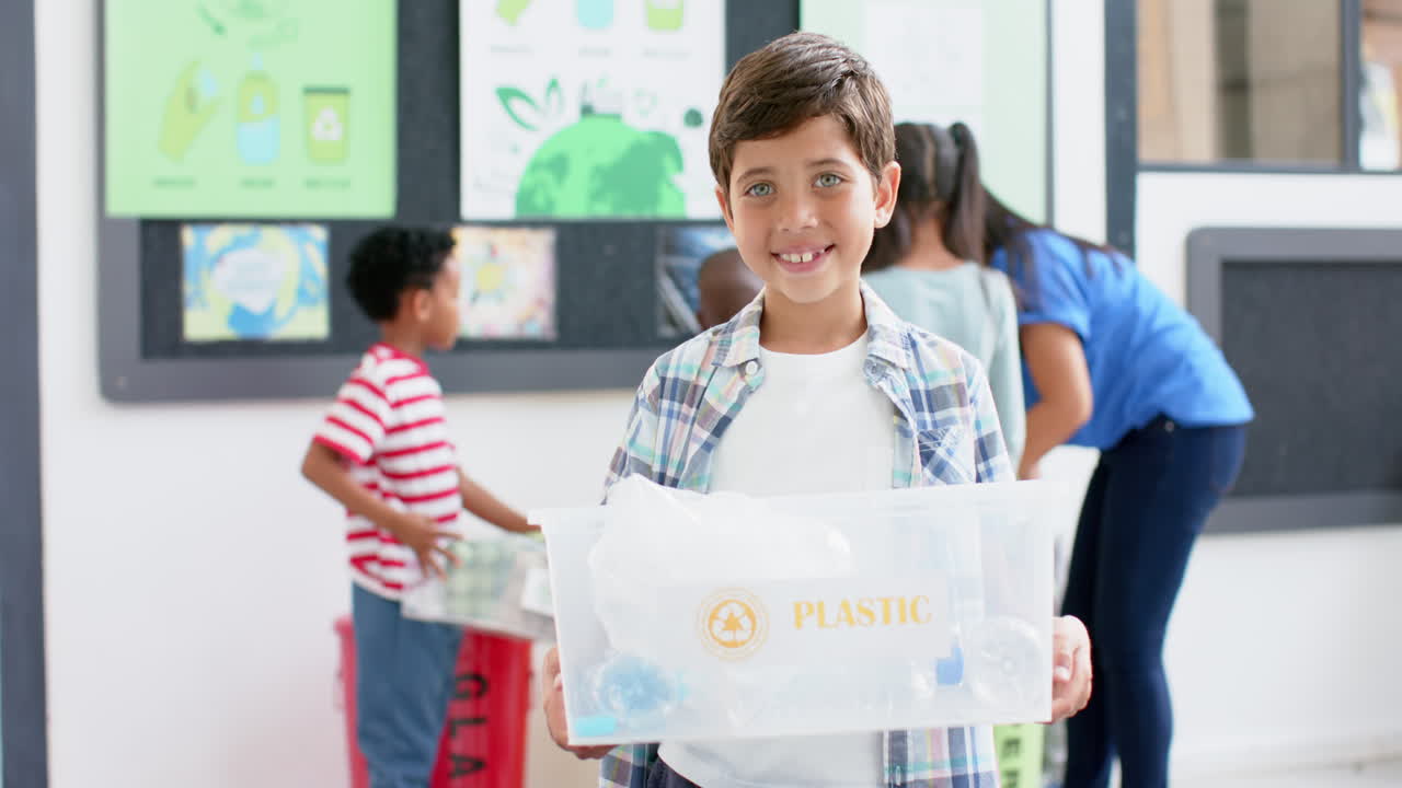 Boy holding plastic recycling bin, smiling in school classroom with classmates