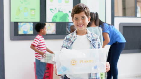 boy holding plastic recycling bin, smiling in school classroom with classmates