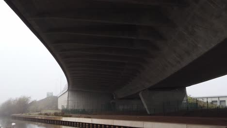 Debajo-De-Una-Gran-Infraestructura-De-Autopista-De-Hormigón-Curvo,-Incline-Hacia-Abajo-Hasta-El-Paisaje-De-Paso-Elevado-Del-Canal-Siniestro-Y-Brumoso