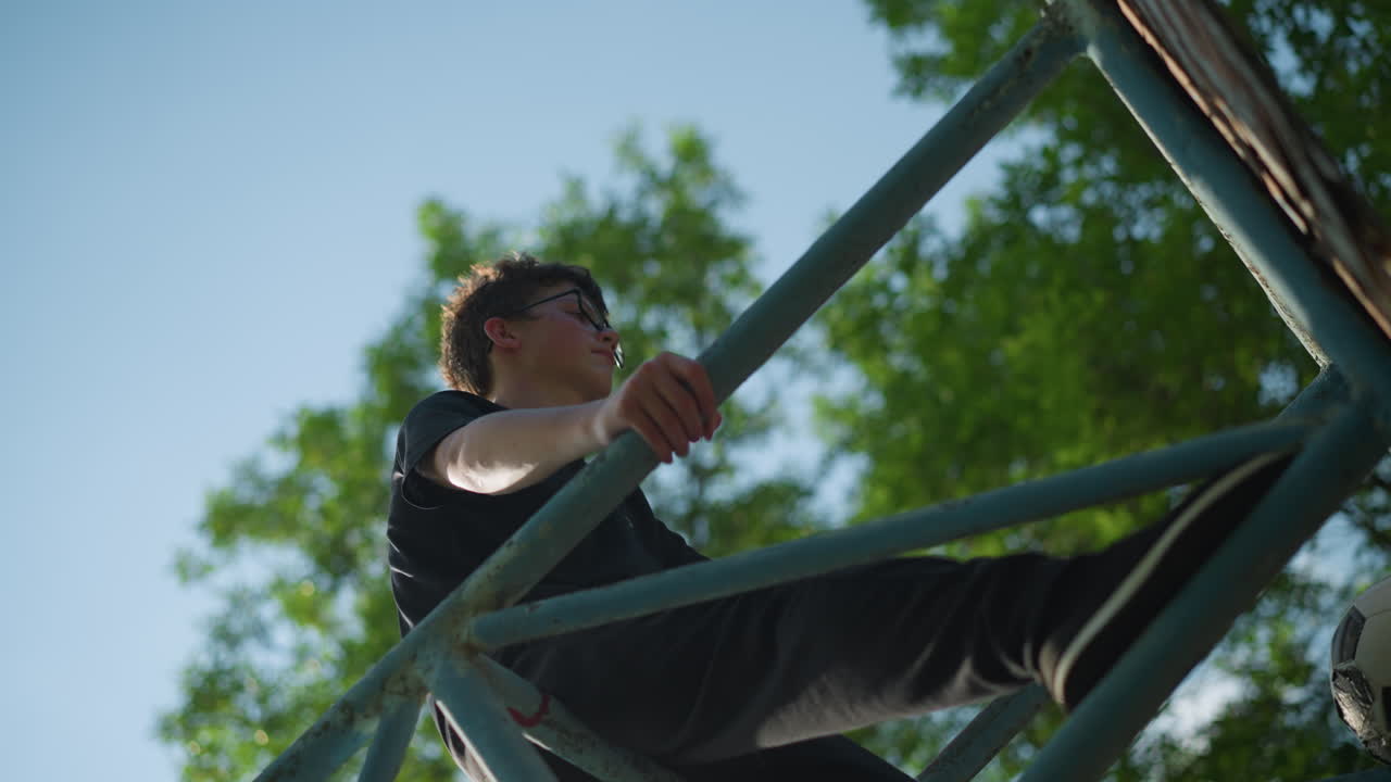 un niño joven está de pie en una estructura de hierro azul al aire libre, mirando pensativo con ambas manos agarrando el marco, una pelota de fútbol desgastada está atascada dentro de las barras de hierro
