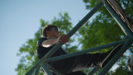 a young boy stands on an outdoor blue iron structure, looking thoughtful with both hands gripping the framework, a worn soccer ball is stuck within the iron bars