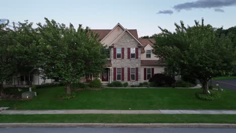 aerial approaching shot of brick single family house with green yard and green waving trees. sidewalk with fire hydrant at clear sky in the morning.