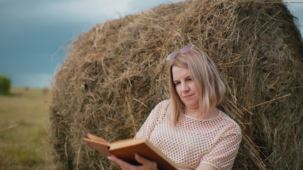 joven con gafas de sol en la cabeza descansando contra la bala de heno leyendo, volviendo a una nueva página, entorno rural sereno con árboles borrosos en el fondo lejano