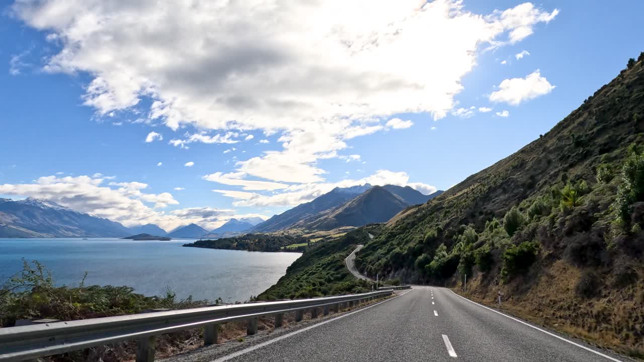 A picturesque drive along a winding road beside Lake Wakatipu, showcasing stunning mountain views under a partly cloudy sky