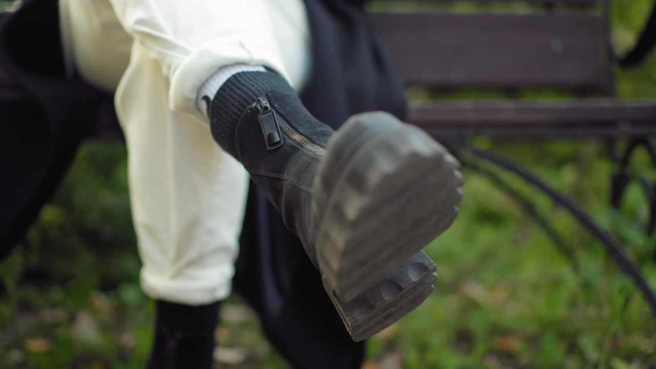 Leg view of serene lady seated on bench with legs crossed, black boots rolling gently, sole toward camera, relaxed motion in autumn park with green trees, soft daylight, quiet mood, casual style