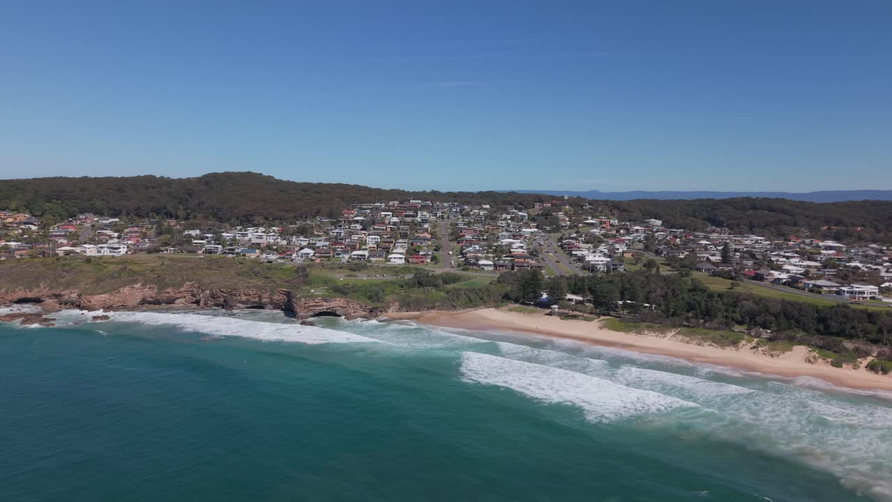 Panoramic aerial establishing of coastal suburb looking over calm water with soft surf and houses perched along cliffs and beachfront roads