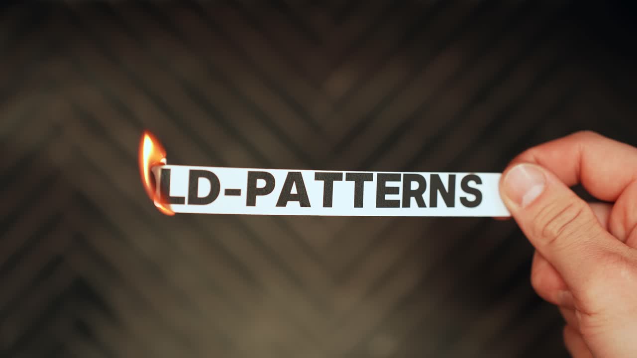 Close-up of a hand holding a paper strip reading “OLD-PATTERNS” while a lighter flame burns the edge, symbolizing breaking habits, personal change, and letting go