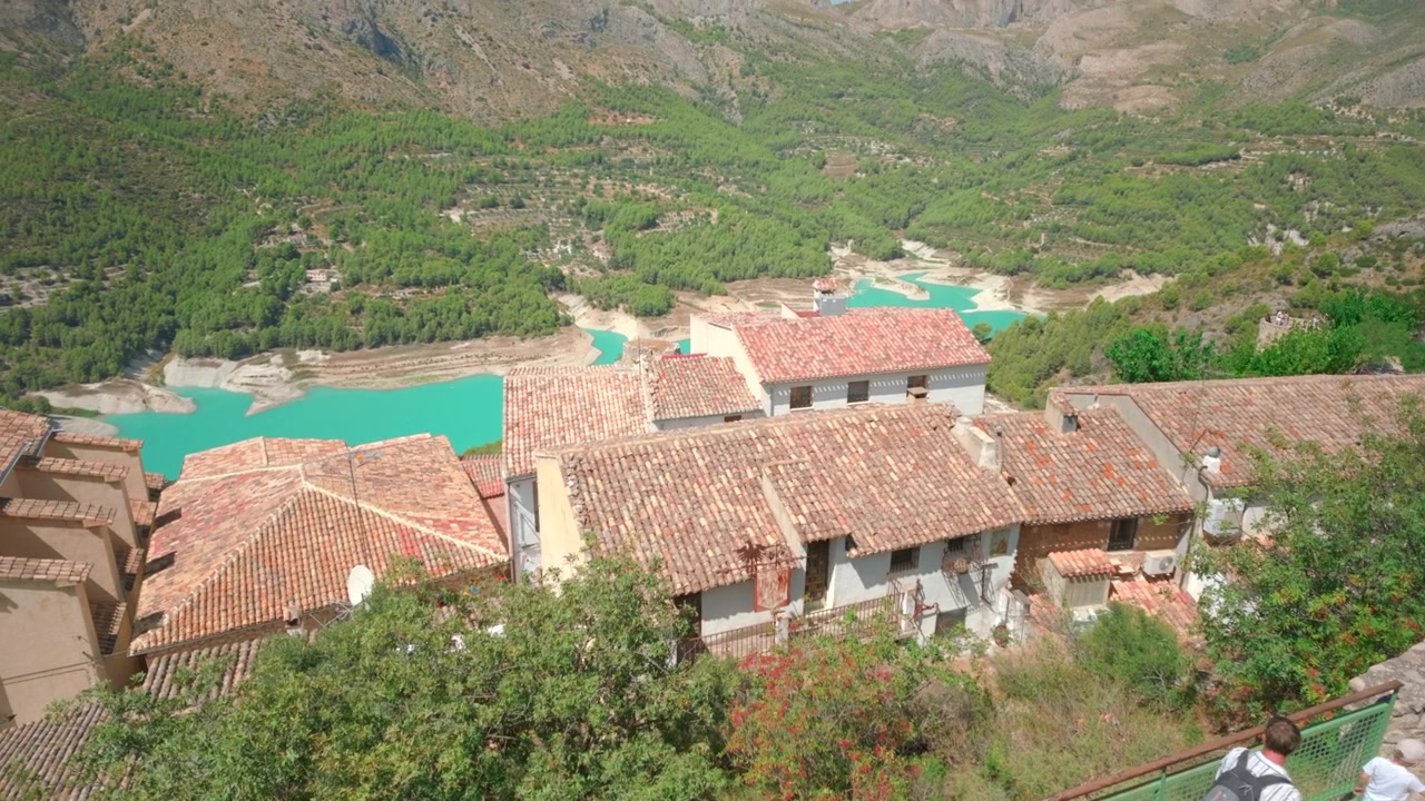 Vista de ángulo alto sobre tejados de terracota, paredes blancas de pueblo y agua turquesa debajo, con densas colinas y montañas verdes bajo la luz brillante del día, pequeños visitantes en la barandilla de la ladera y calma.