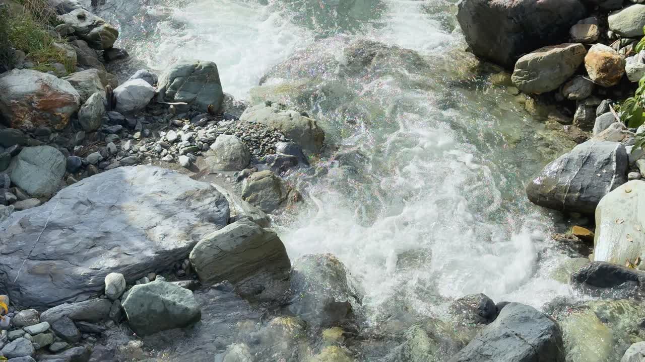 Clear glacial meltwater rushes over smooth rocks in a sunlit mountain stream. Static camera captures natural movement, crisp daylight, and vibrant textures
