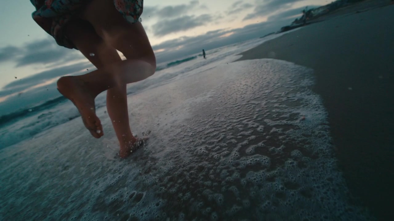 Un'energica inquadratura dal basso segue una donna a piedi nudi che corre tra le onde basse su una spiaggia sabbiosa al crepuscolo dorato, con gli spruzzi del mare che volano, pura gioia, libertà e vitalità estiva.