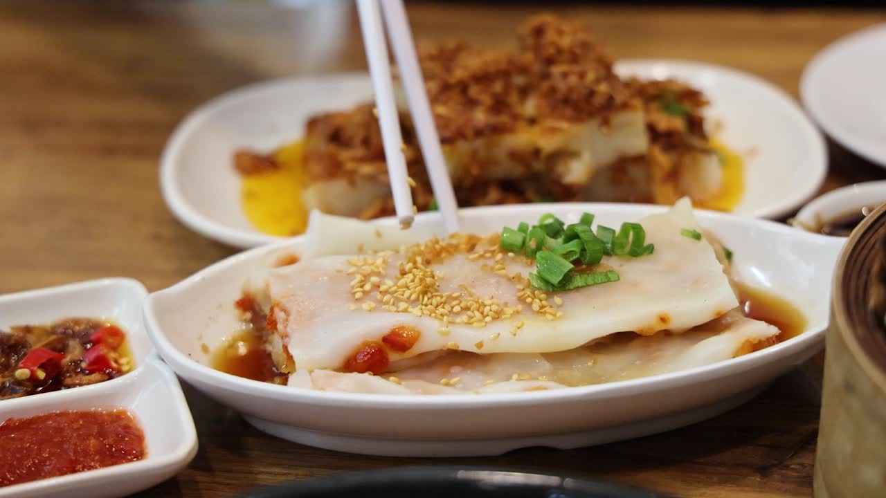 Chopsticks grasp and lift steamed rice noodle rolls garnished with scallions and sesame seeds, set on a wooden table with side dishes in warm, natural lighting