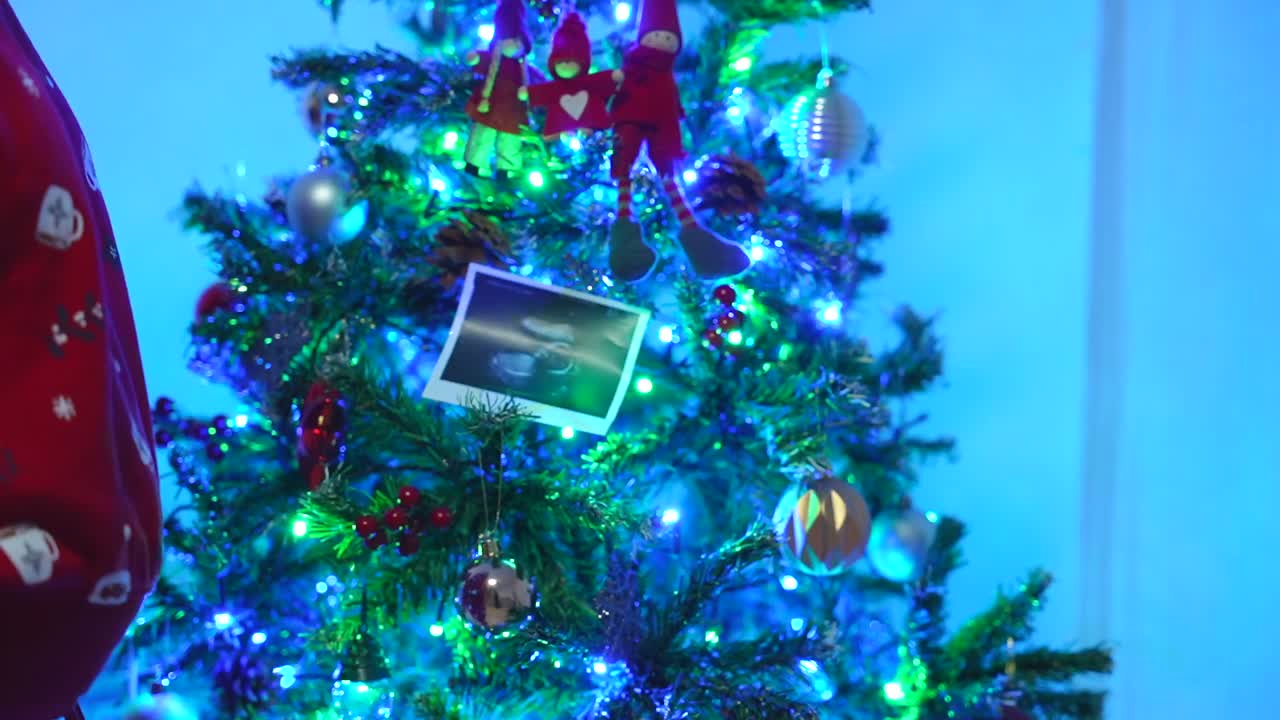 Pregnant woman in red pajamas places an ultrasound image on the illuminated Christmas tree while tenderly touching her exposed belly