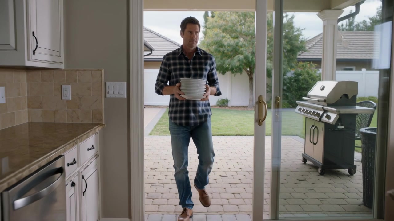 Walking plaid man holding white bowls into kitchen via open sliding glass door to load dishwasher
