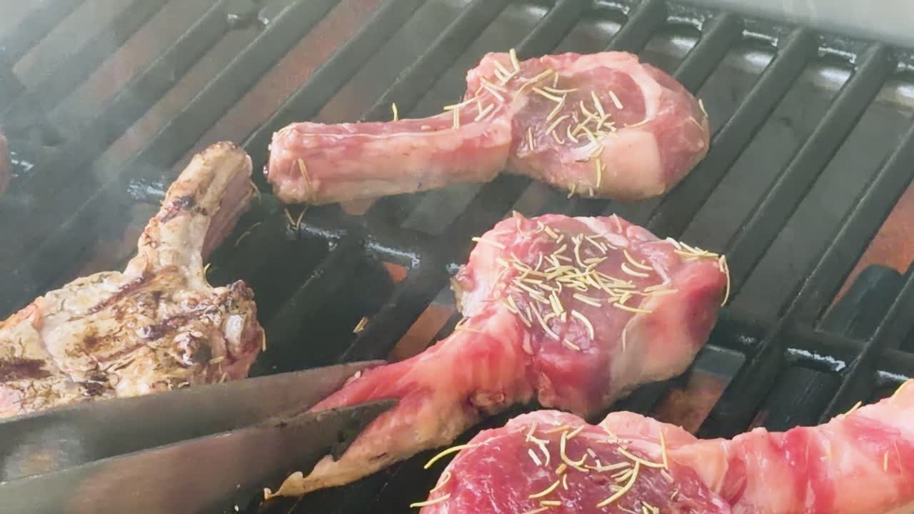 Lamb cutlets cook over open flame, flipped with tongs, producing smoke and grill marks