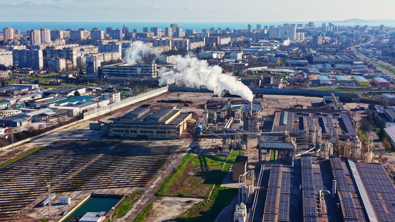 Industrial area with factories and pollution near a city skyline by the coast during the daytime with smoke and solar panels visible