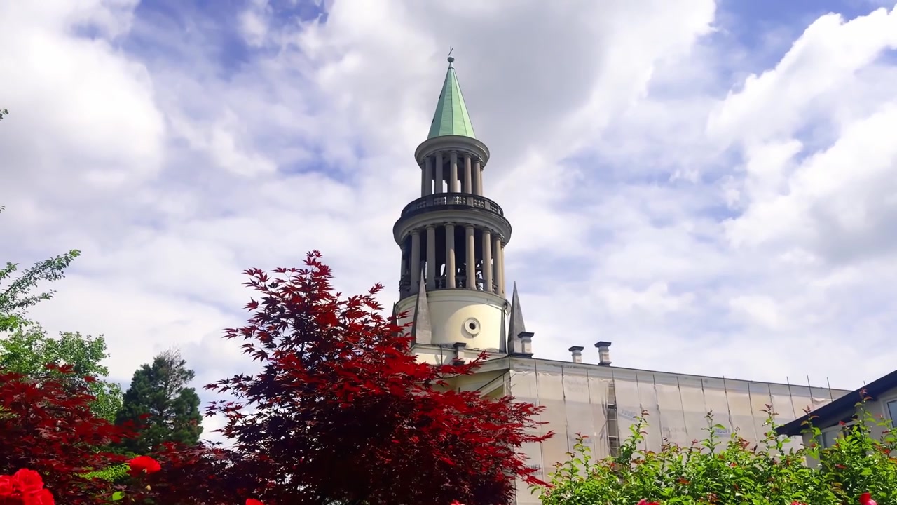 Sankt Georgs kyrka i Tacen, Ljubljana, reser sig över röda lönnlöv och trädgårdsblommor under ljusa sommarmoln.