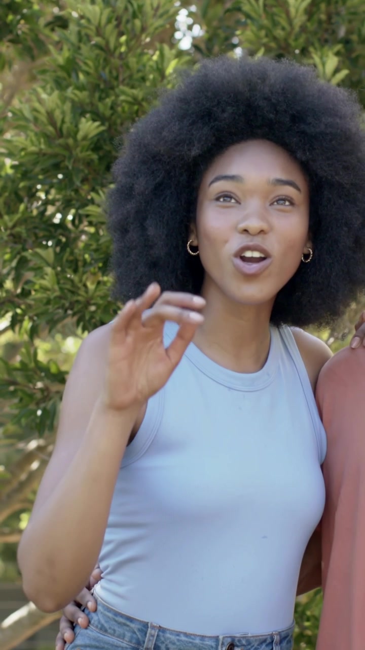Vertical video: Smiling woman in blue top lifting hand in garden, reacting off-frame, hand on waist
