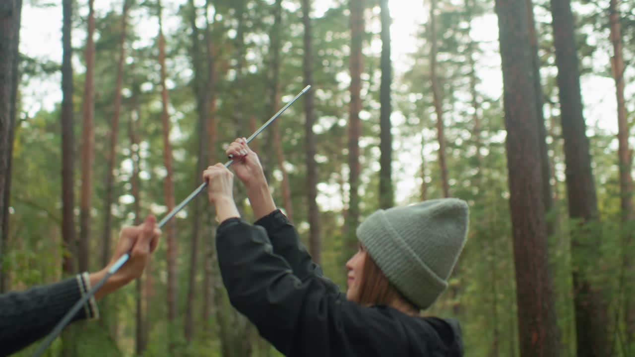 Young lady with warm smile holds tent pole while man assists her in connecting pieces for camping setup in tranquil forest, enjoying shared moment of teamwork surrounded by tall trees and fresh air