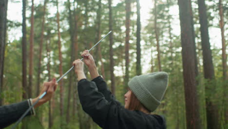 young lady with warm smile holds tent pole while man assists her in connecting pieces for camping setup in tranquil forest, enjoying shared moment of teamwork surrounded by tall trees and fresh air