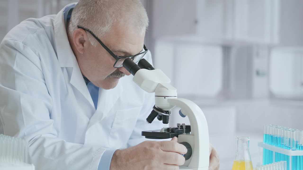 Bearded Caucasian male researcher wearing protective glasses and working with a microscope spbas. scientist using microscope in a laboratory. Search for coronavirus vaccine. High quality 4k footage
