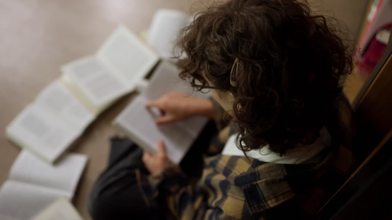 más de la vista superior del hombro de una estudiante con el cabello rizado usando gafas sentado en el suelo y leyendo un libro en la biblioteca