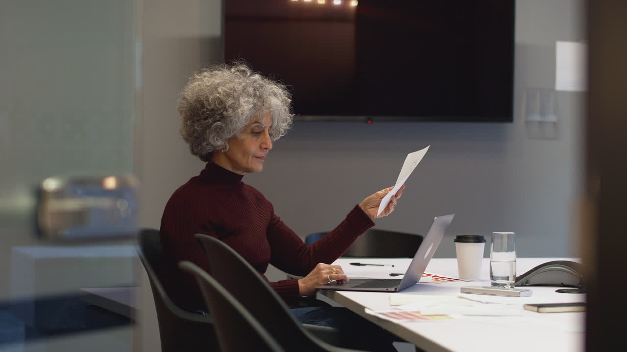 Donna d'affari matura che lavora fino a tardi in una sala riunioni dell'ufficio utilizzando il laptop