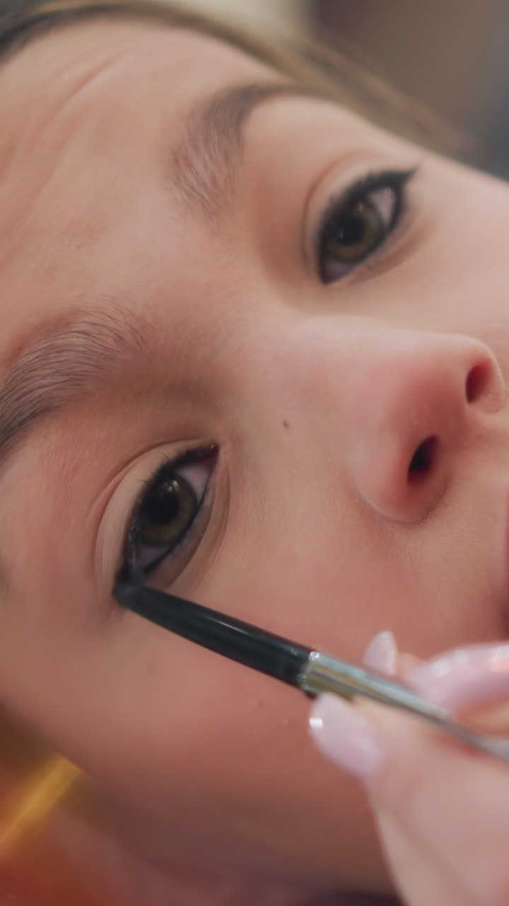 Primer plano frontal de una mujer sin especificar delineando las pestañas inferiores con un pincel pequeño, ambiente íntimo de tutorial de belleza y pulso firme, influencer ensayando y preparando la definición de los ojos con trazos cuidadosos, luz natural