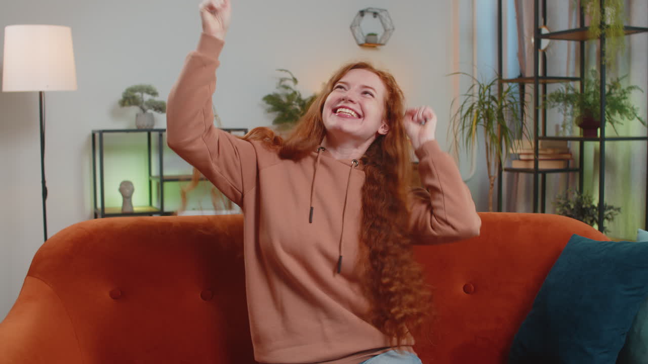 Happy young woman watching tv celebrating success winning lottery luck victory sitting on sofa