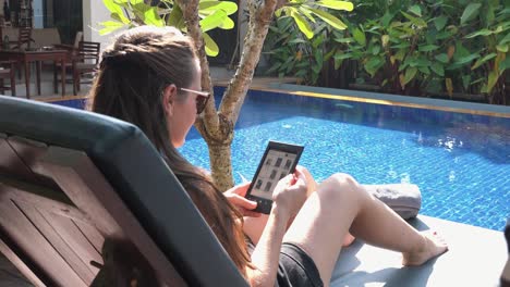 tiro medio por encima del hombro de una mujer joven leyendo un libro electrónico en una tableta junto a la piscina