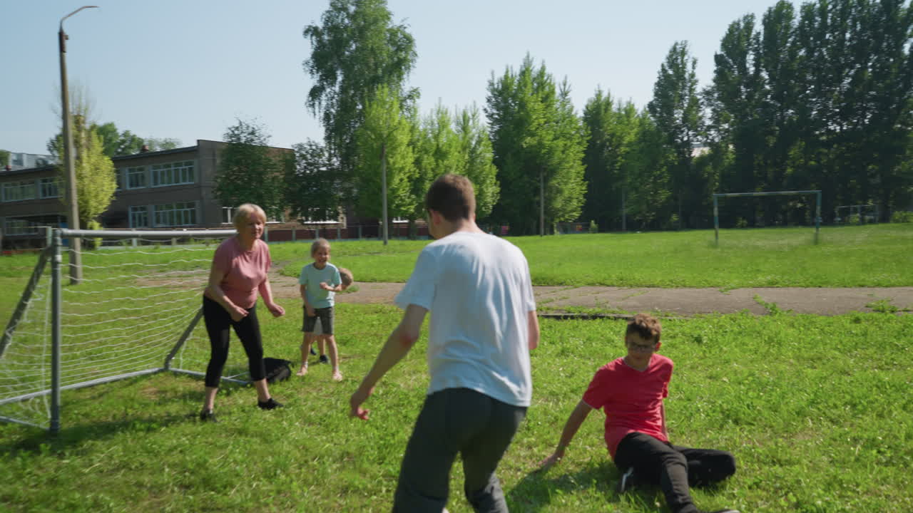 la familia disfruta de un animado juego de fútbol al aire libre mientras el hijo vestido de blanco dribla la pelota más allá de su hermano deslizante y anota en el poste de la portería, con su madre viendo la pelota entrar en la red