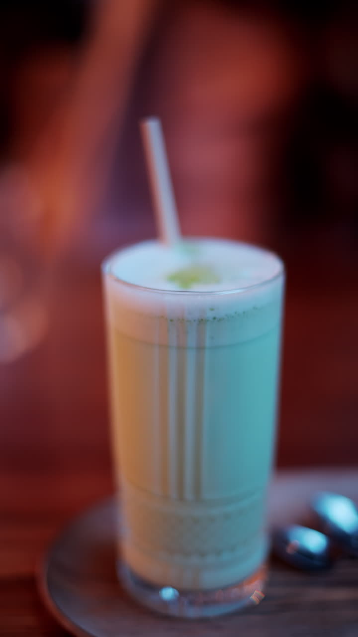 Close up of a matcha latte in a glass with a straw on a table at a cafe. Vertical