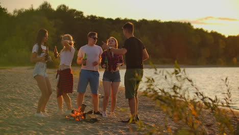 un grupo de cinco jóvenes están bailando en pantalones cortos y camisetas alrededor de una fogata en la naturaleza con cerveza. están haciendo clink y bebiendo cerveza y disfrutando de la tarde calurosa al atardecer cerca del lago.