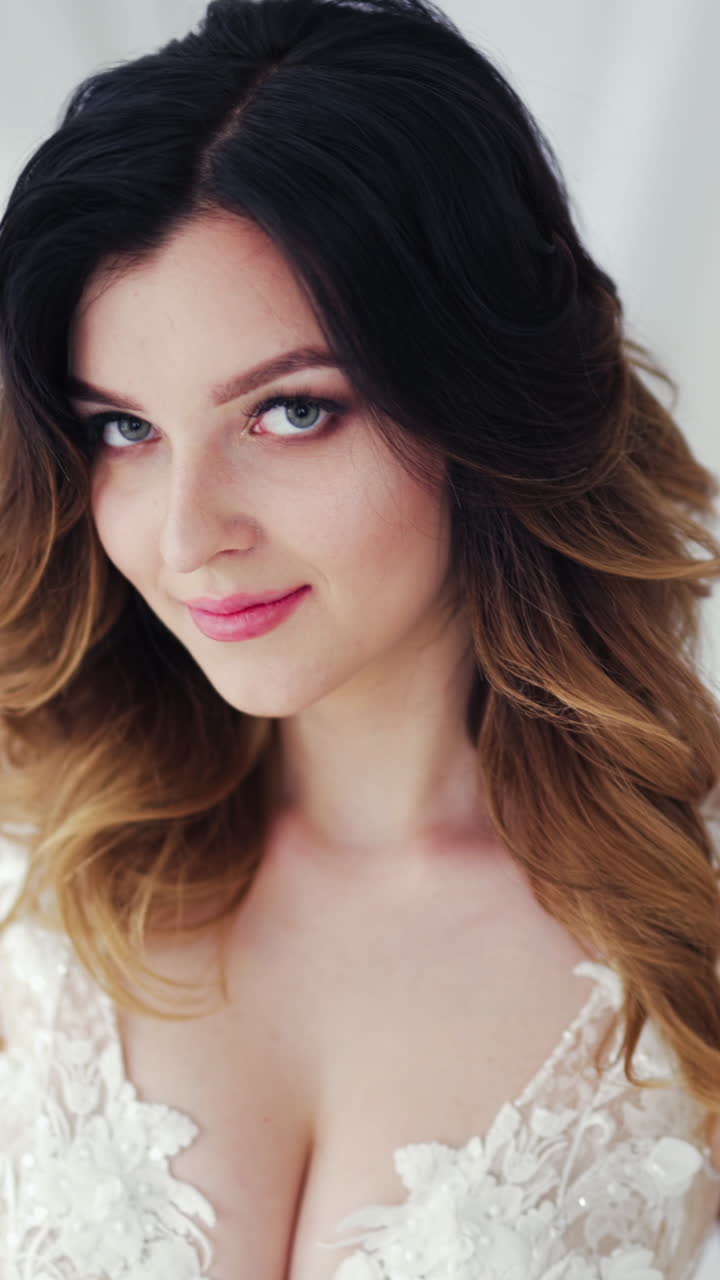 Portrait of a beautiful young bride with wavy hairdo smiling into camera. Glamour model with stylish makeup is sitting on bed and posing in the studio. Vertical video