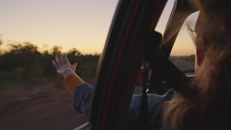 woman in car holding hand out window feeling wind blowing through fingers driving in countryside on road trip travelling for summer vacation enjoying freedom on the road at sunrise