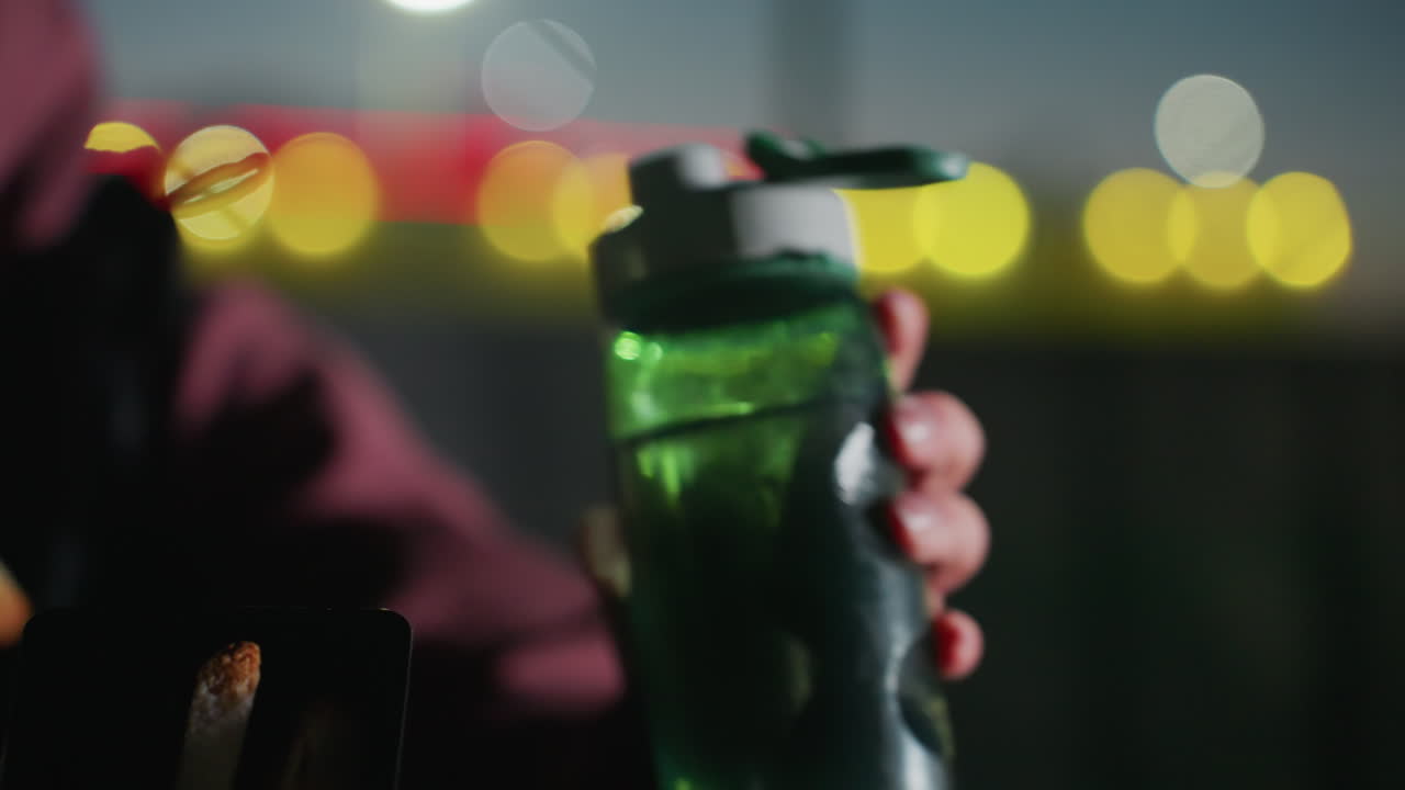 Close up of athlete hand grabbing chilled water bottle for refreshing sip then placing back on wooden picnic bench under blurred city lights at twilight in urban park for hydration break