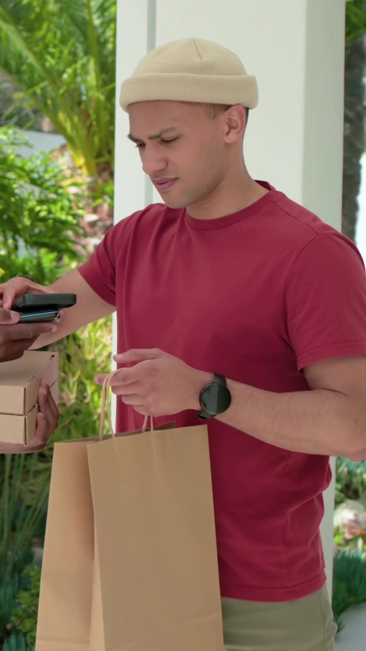 Vertical video: Receiving boxes, man red tee checking phone for pickup at porch, stacking brown bag