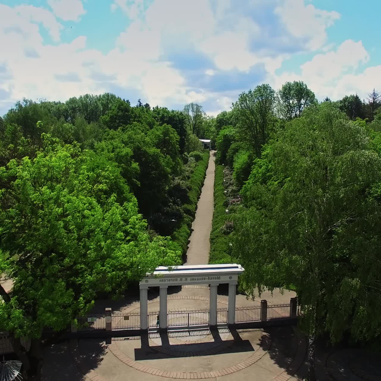 Flying closer to the city park with white columns at the entrance. Long path through the green trees leading to the building. Top view