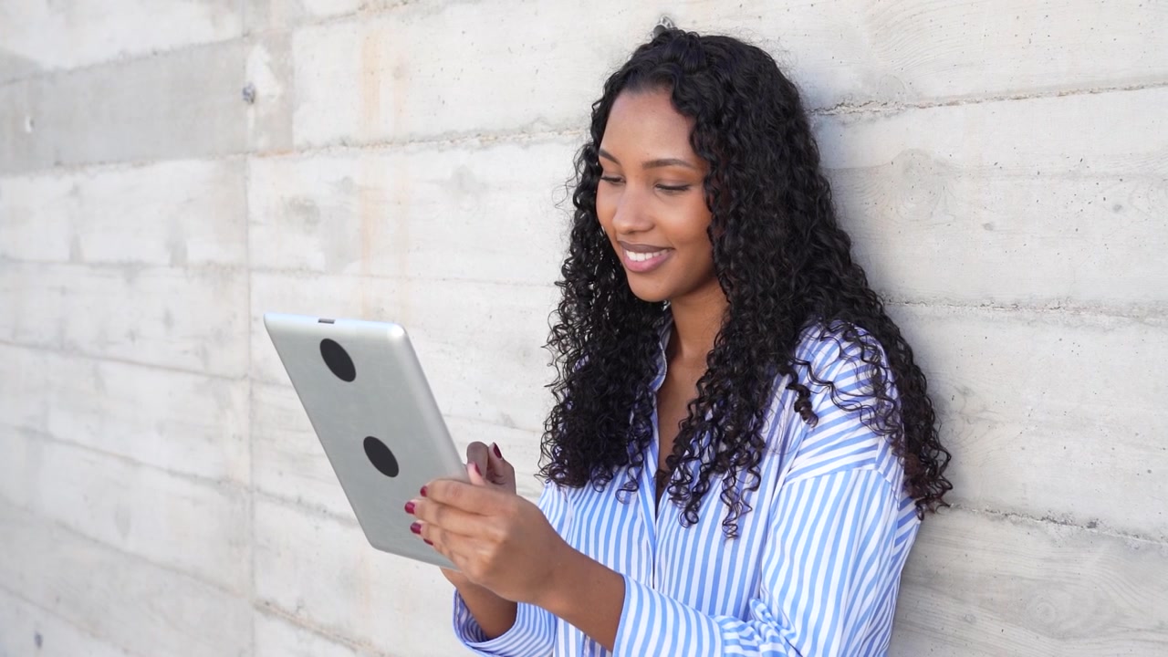 A focused woman deeply engaged while using a tablet leaning against a concrete wall