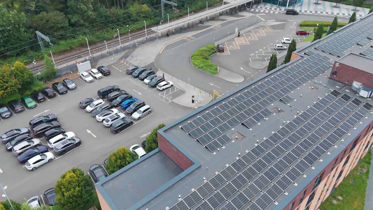 Widok z lotu ptaka na budynek z panelami słonecznymi na dachu, otoczony parkingiem i zielenią. Zakrzywiona droga i zaparkowane samochody podkreślają połączenie infrastruktury miejskiej i zrównoważonego projektowania.