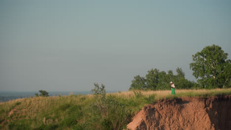 distant woman in white hat and green skirt approaches cliff edge holding camera while standing amid golden grass and lush trees under clear blue sky capturing scenic landscape on sunny day