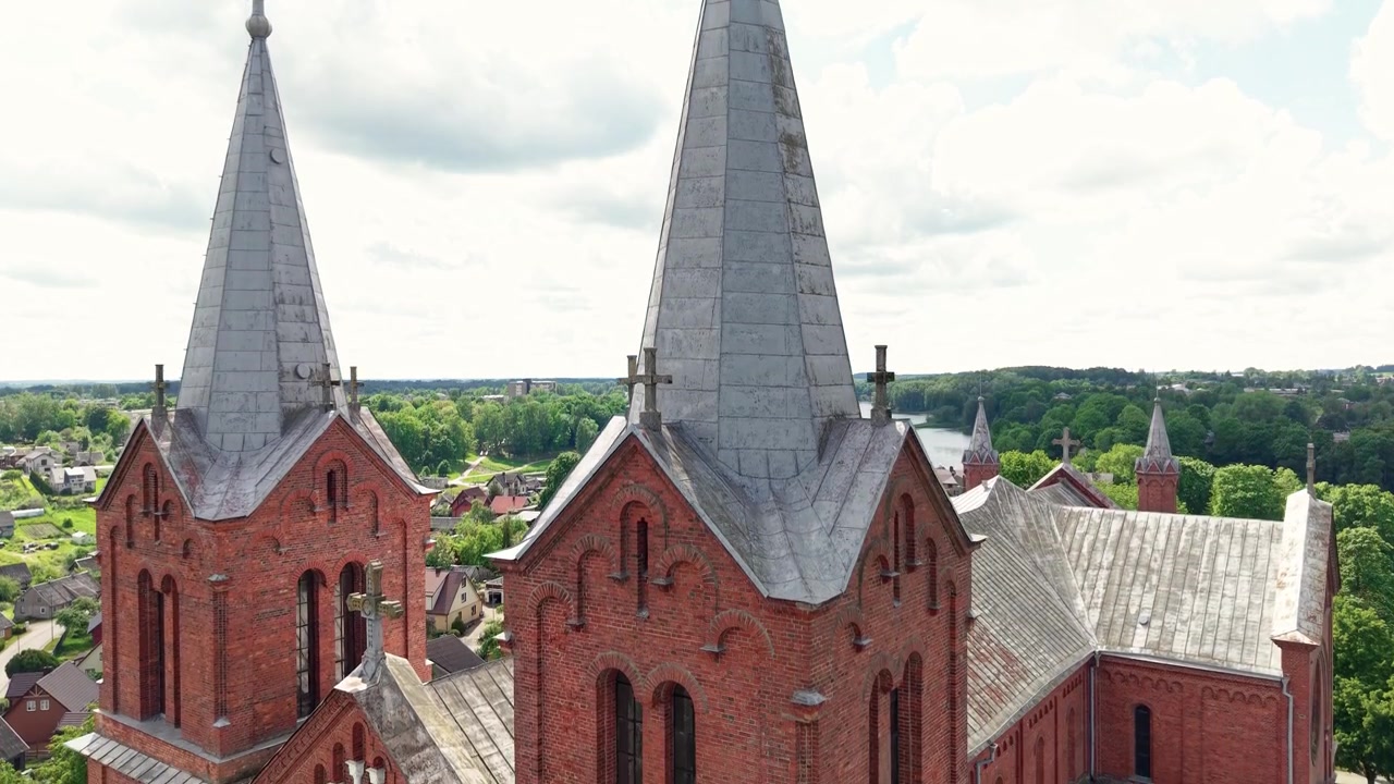 Majestätisk flygfotografering med drönare glider över Johannes Döparens kyrka i Plungė, Litauen, och avslöjar utsmyckade spiror, frodig grönska, fridfulla tak och en glittrande flod under starkt dagsljus.