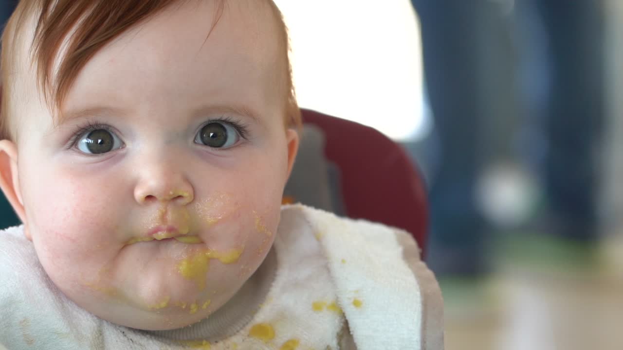 A mother's hand scooping up baby food off of a toddlers face with a spoon, Front facing shot - slow motion