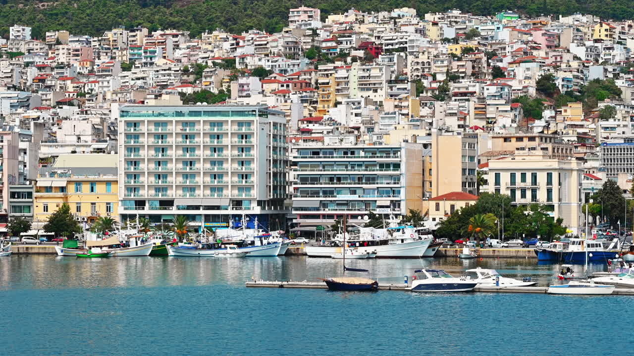 KAVALA, GREECE - SEPTEMBER 23, 2020: Multiple buildings on the Aegean sea coast with port and moored yachts, green hill on the background