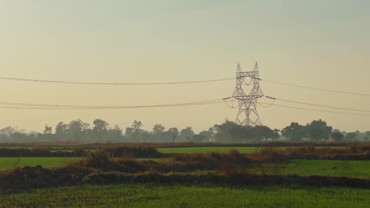Long shot of a high voltage transmission tower standing over green agricultural fields at evening light, power lines stretching across horizon with trees and calm rural landscape